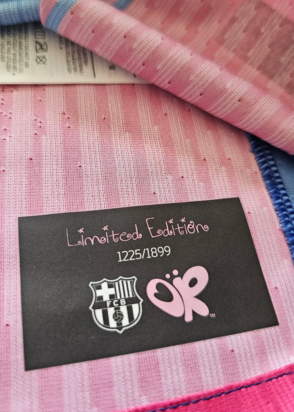 Black 'Limited Edition 1225/1899' tag with the FC Barcelona crest and pink Olivia Rodrigo butterfly logo, sewn into Frenkie de Jong's shirt.