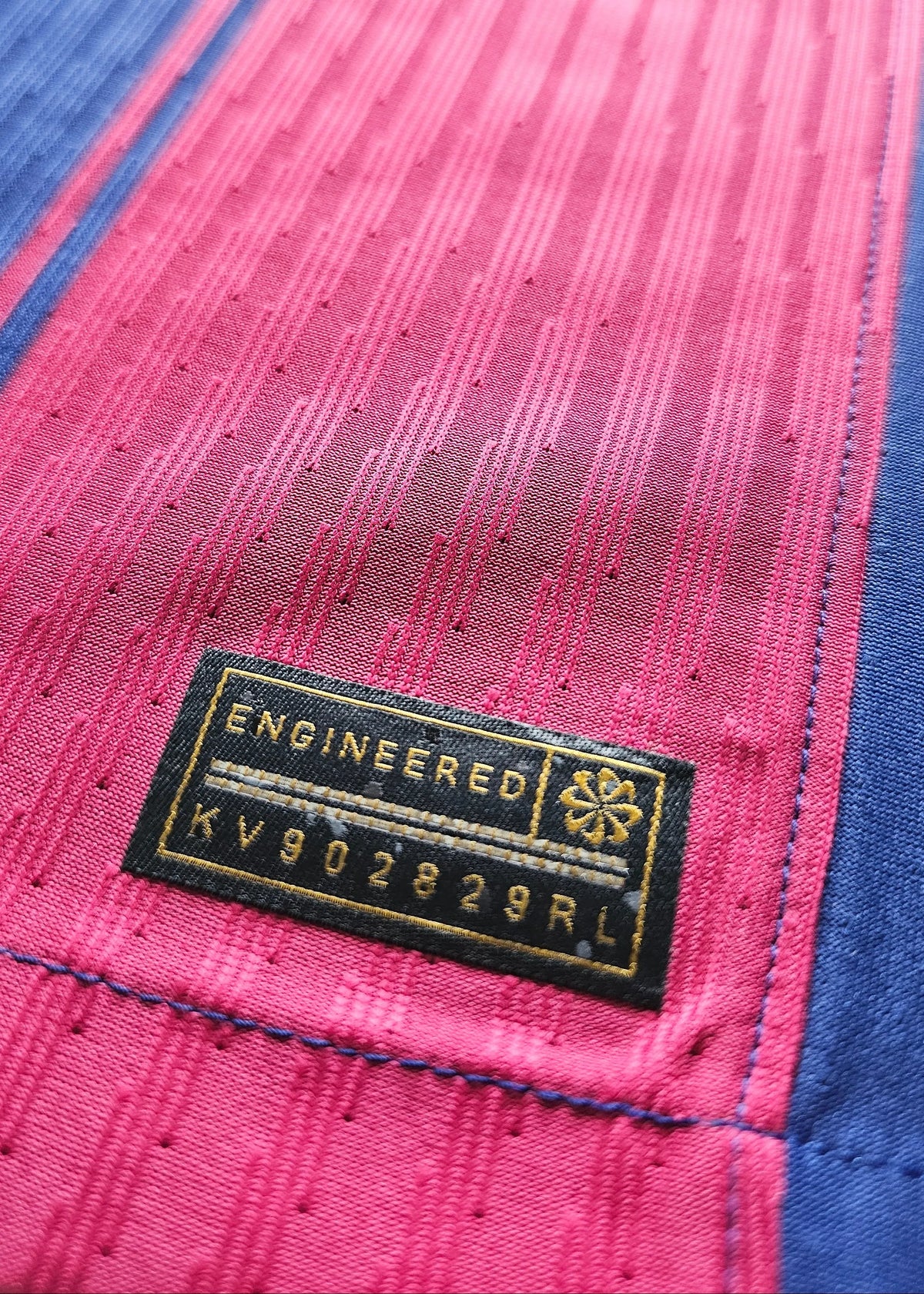 Black woven Nike 'Engineered' tag with a small gold emblem and style code KV902829RL, stitched onto the pink fabric of Frenkie de Jong's shirt.