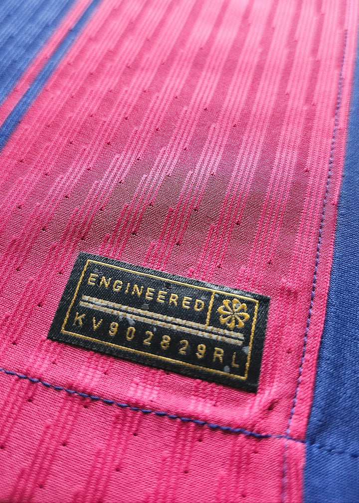 Black woven Nike 'Engineered' tag with a small gold emblem and style code KV902829RL, stitched onto the pink fabric of Frenkie de Jong's shirt.
