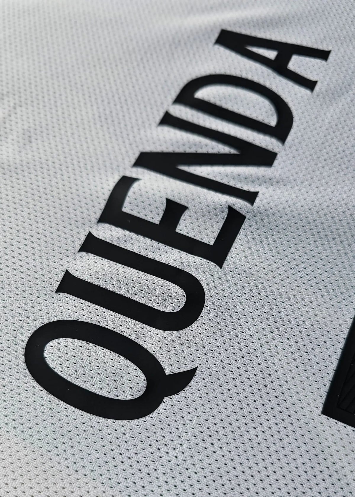 Close-up of a white athletic jersey with black perforated lettering reading 'QUENDA' on the back.