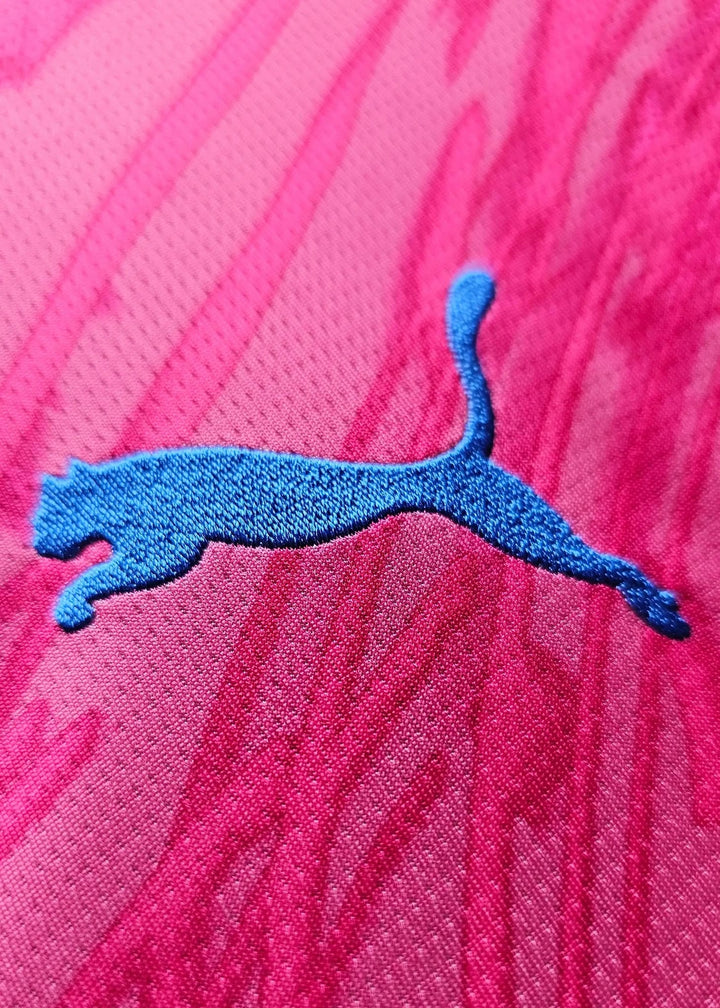 Close-up of the embroidered teal Puma cat logo on the pink fabric of a signed Manchester City goalkeeper shirt.