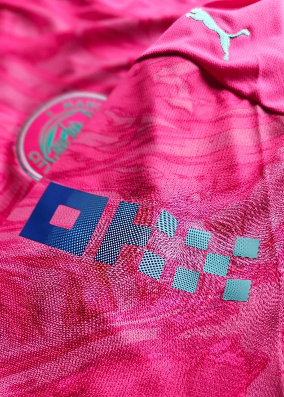 Close-up of the teal Puma cat logo on a pink Manchester City shoulder, with the edge of the club crest visible at left.