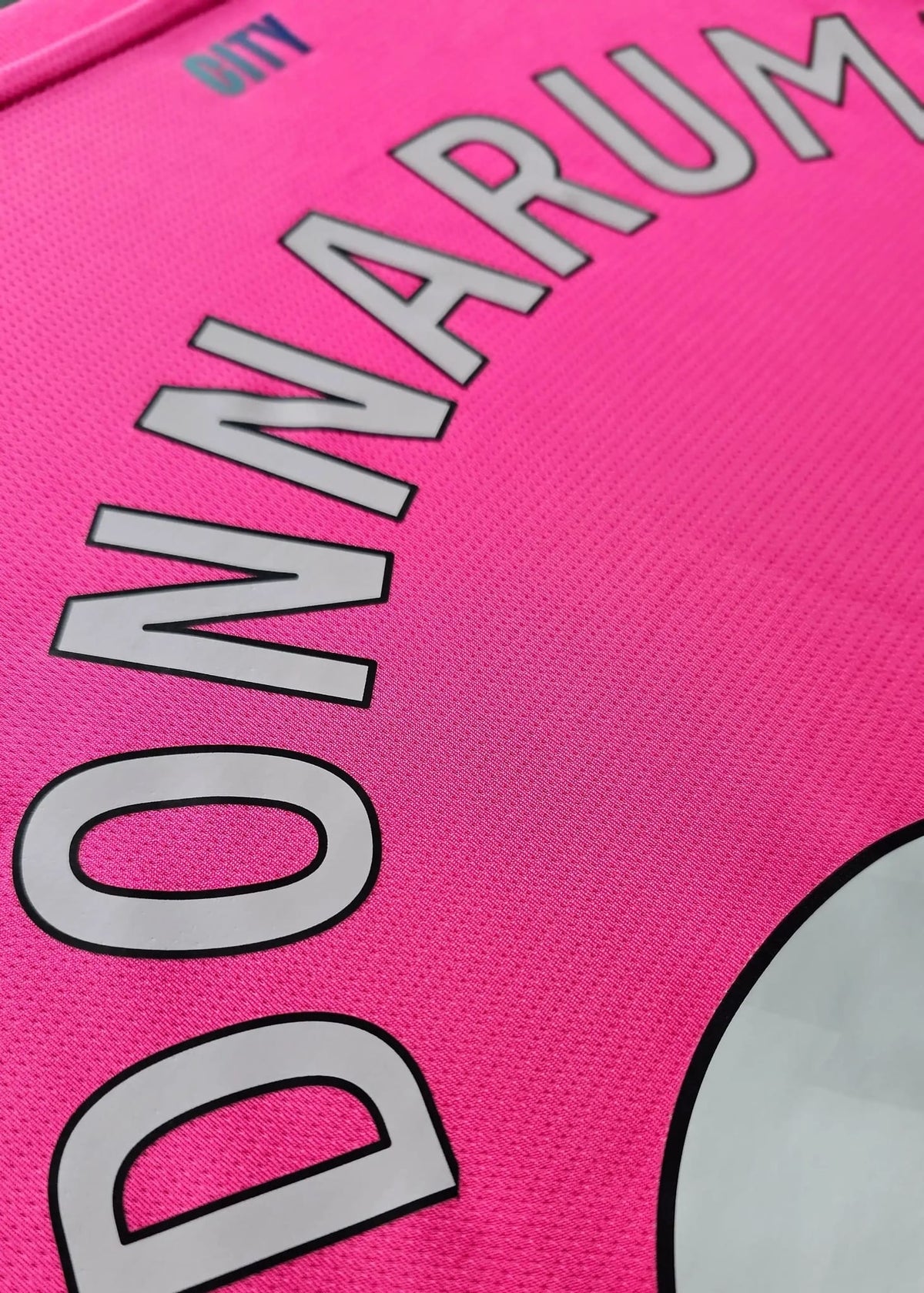 Angled close-up of the printed 'DONNARUMMA' lettering on a pink Manchester City shirt, with a small teal 'CITY' logo above.