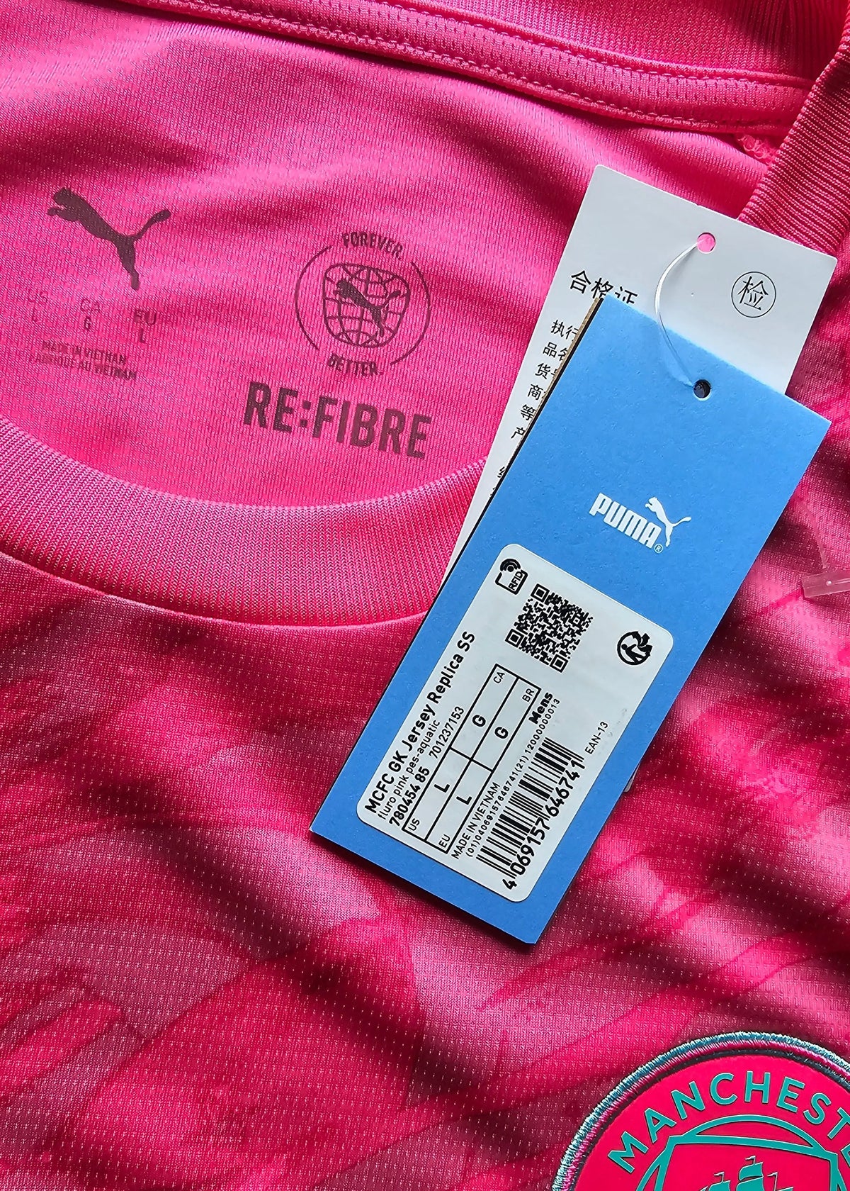 Interior neck of a pink Manchester City shirt with a 'RE:FIBRE' stamp, a hanging Puma size tag, and the edge of the club crest.