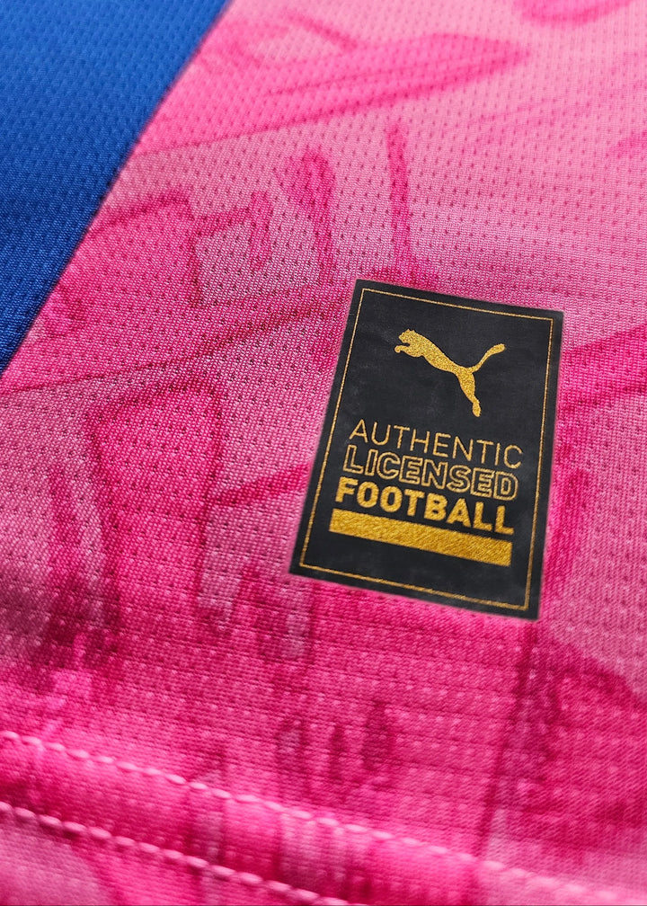 Close-up of a black and gold 'Authentic Licensed Football' Puma patch stitched onto a pink Manchester City shirt near a blue panel.