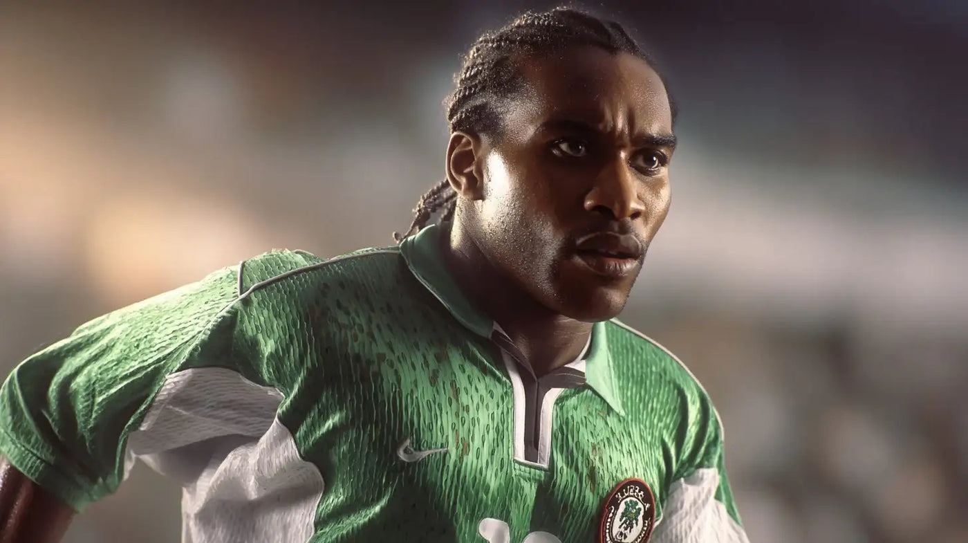 Jay-Jay Okocha wearing a Nigeria 1994 green football shirt with white sleeves and a dark trimmed collar, in match action with a blurred stadium background.
