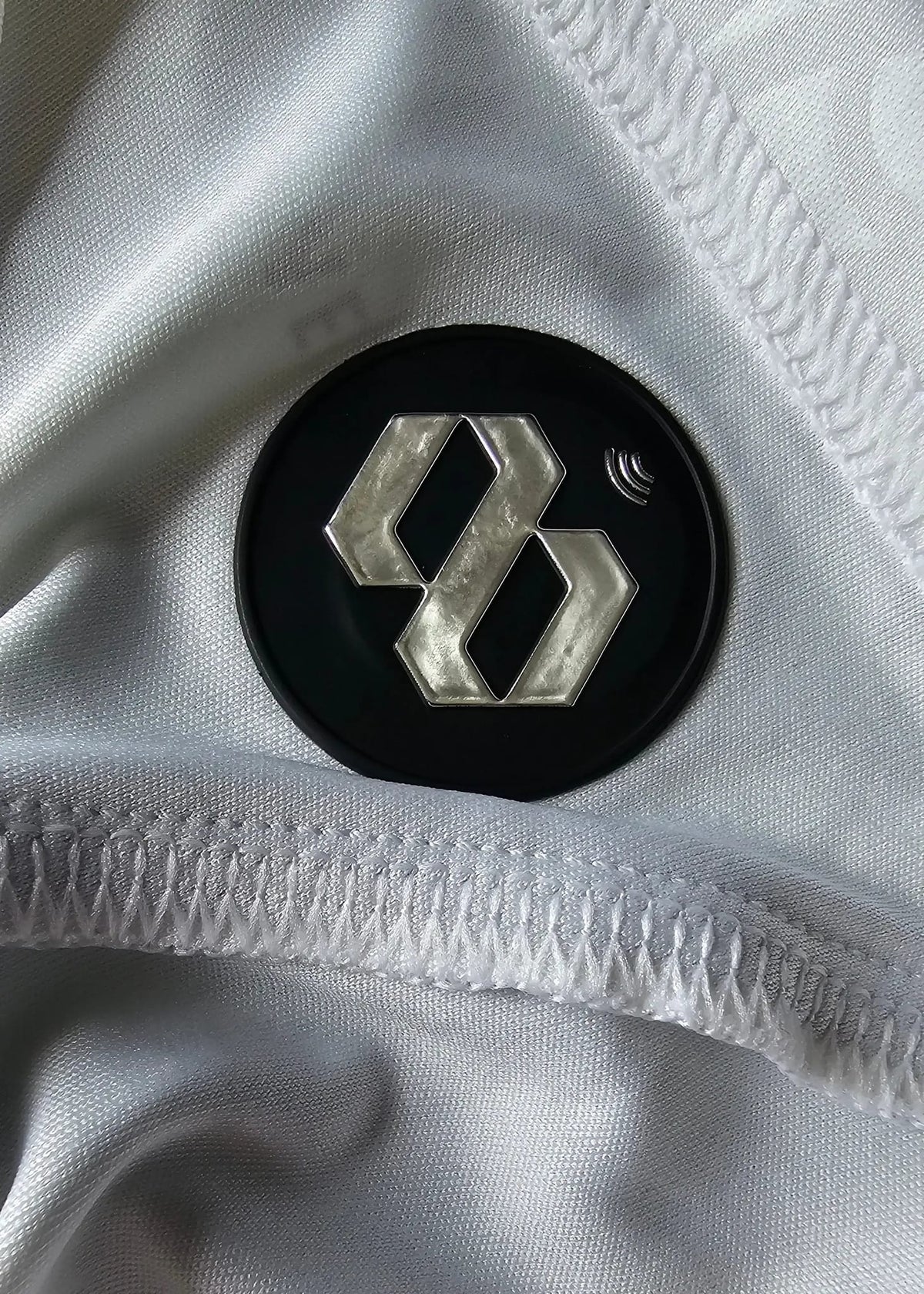 Close-up of a black circular patch with a silver geometric logo sewn onto white textured shirt fabric.