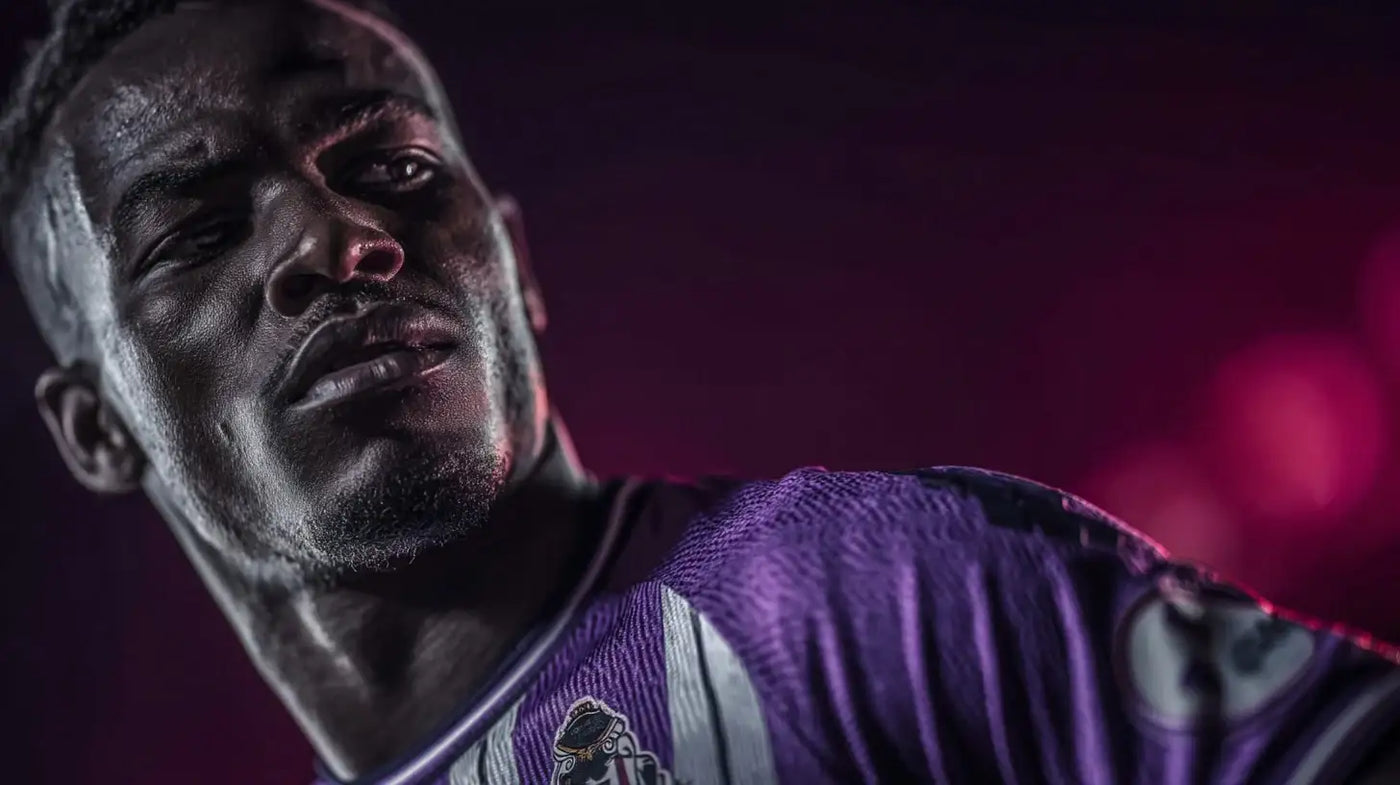 Juma Bah of Real Vailladolid, dramatic side profile in purple-white kit, looking over his shoulder against a pink-lit backdrop.