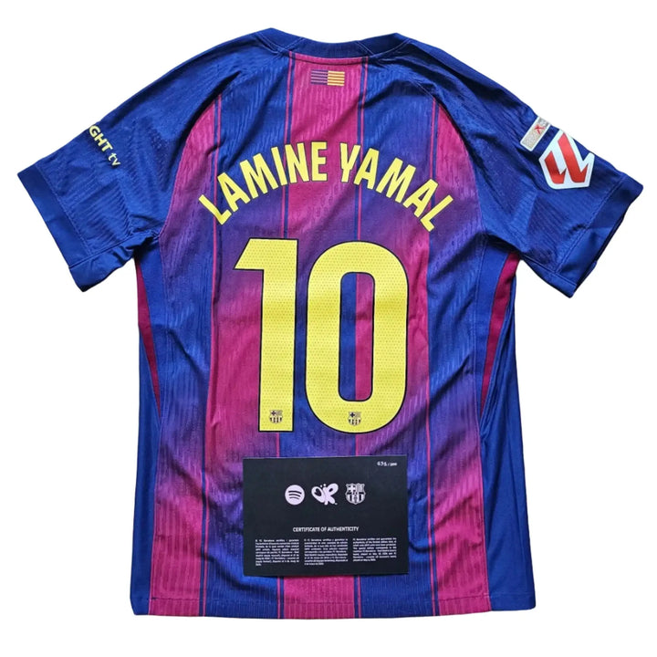 A back view of a Lamine Yamal number 10 FC Barcelona jersey in blue and magenta stripes, laid flat on a light wood floor.