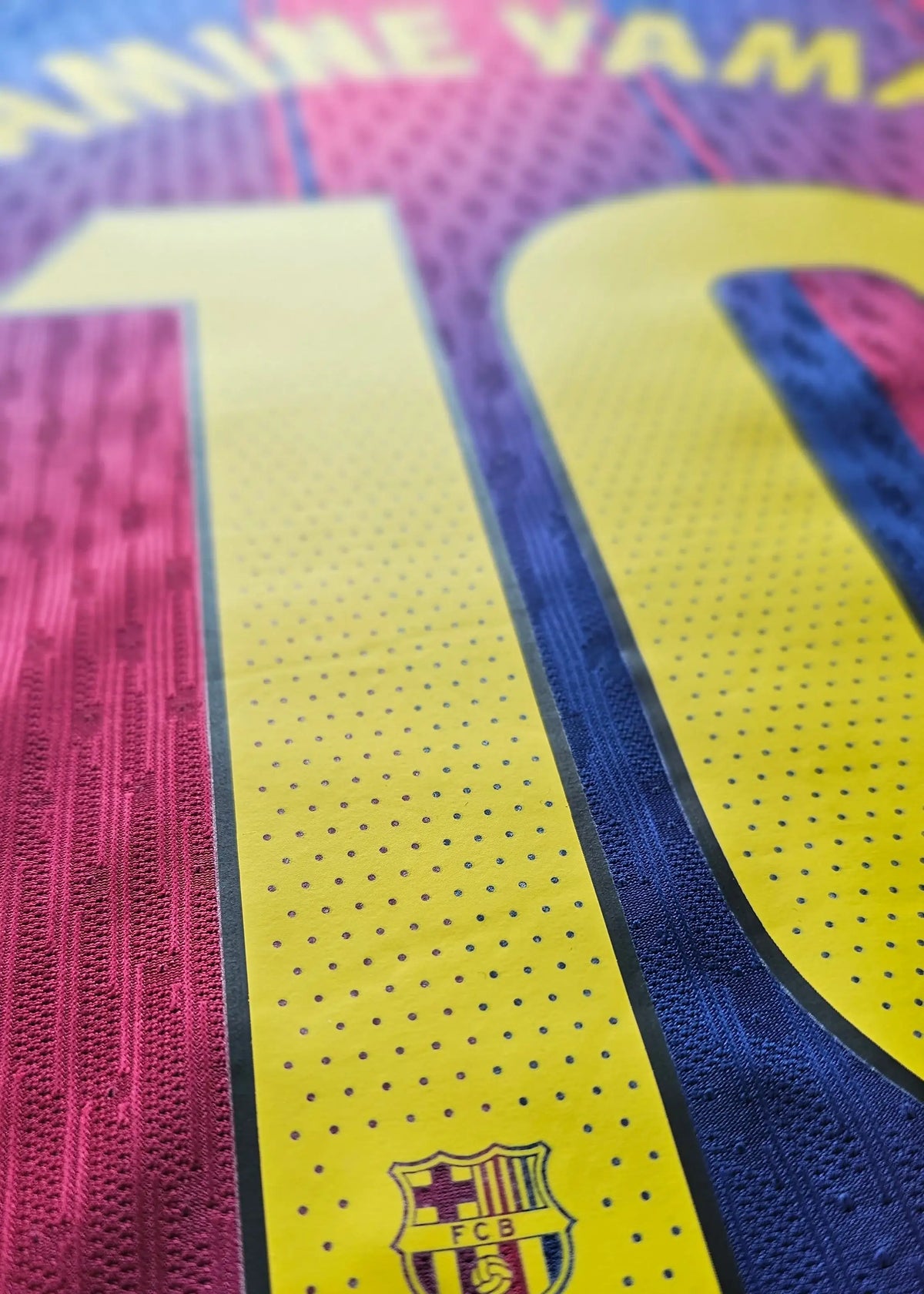 Close-up of a soccer jersey showing the number 10 in yellow with a dotted texture, bordered by pink and blue sections. The FC Barcelona crest is visible near the bottom of the yellow number.