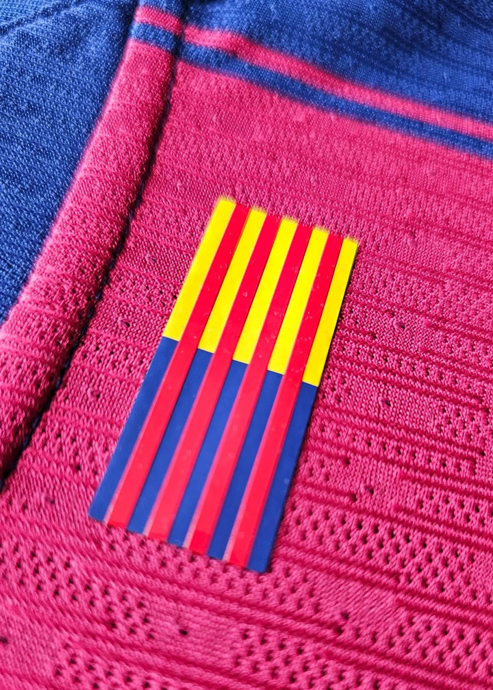 Close-up of the FC Barcelona flag patch in red, yellow, and blue on the pink and blue textured shirt fabric.