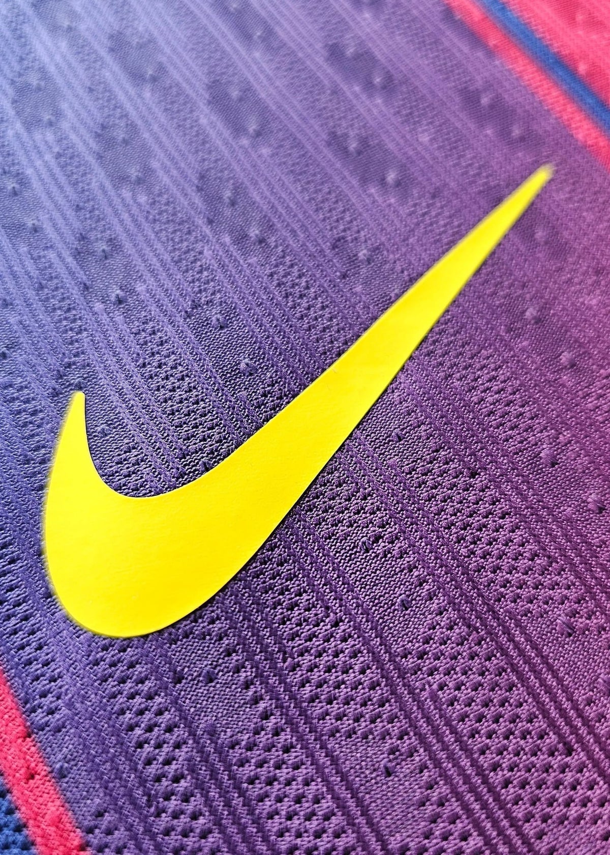 Close-up of a textured purple athletic fabric with a large bright yellow Nike swoosh logo and thin red and blue stripe accents.