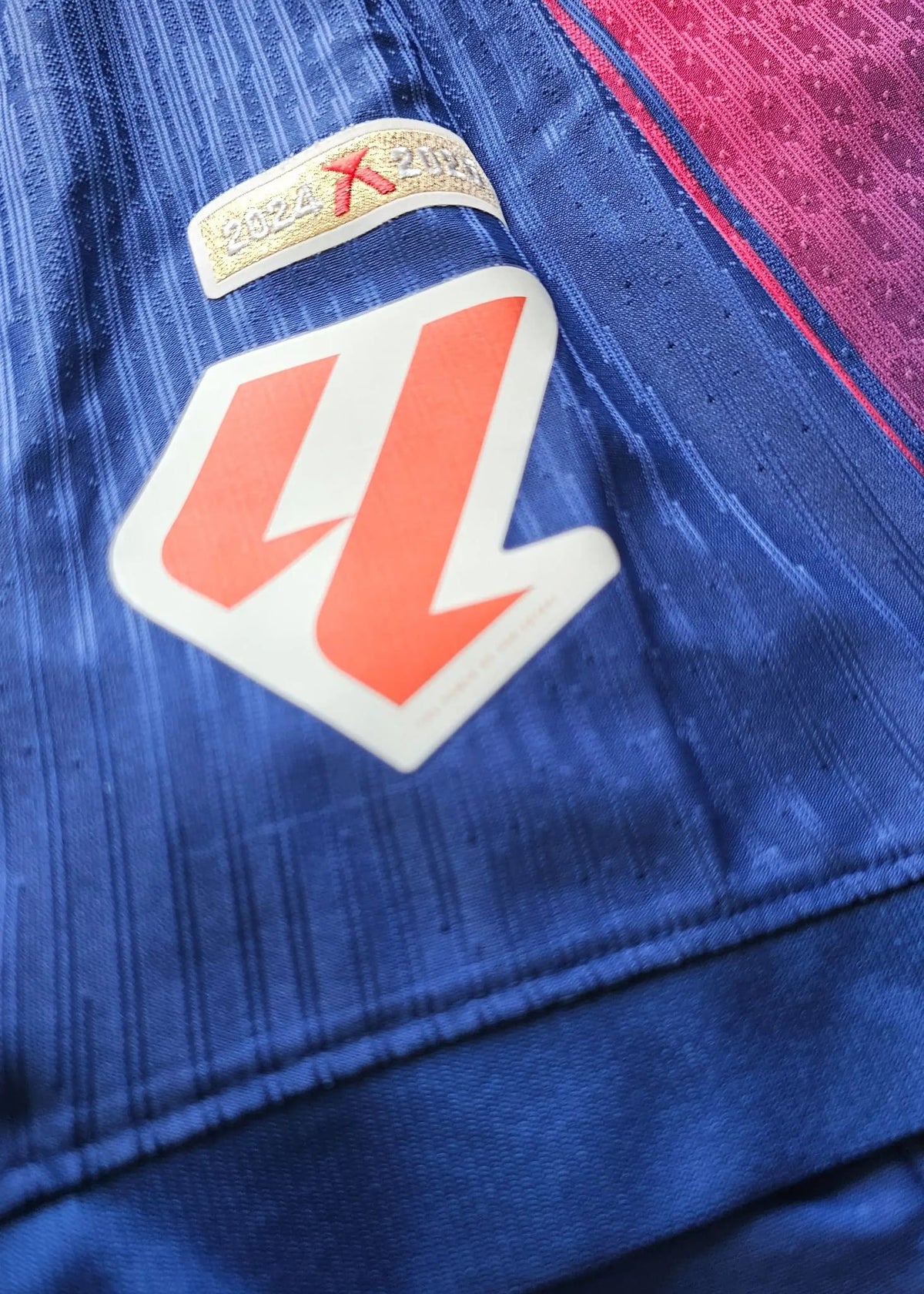 A gold '2024 X 2025' season tab above an orange-and-white angular badge patch, stitched onto the royal blue sleeve fabric of the FC Barcelona shirt.