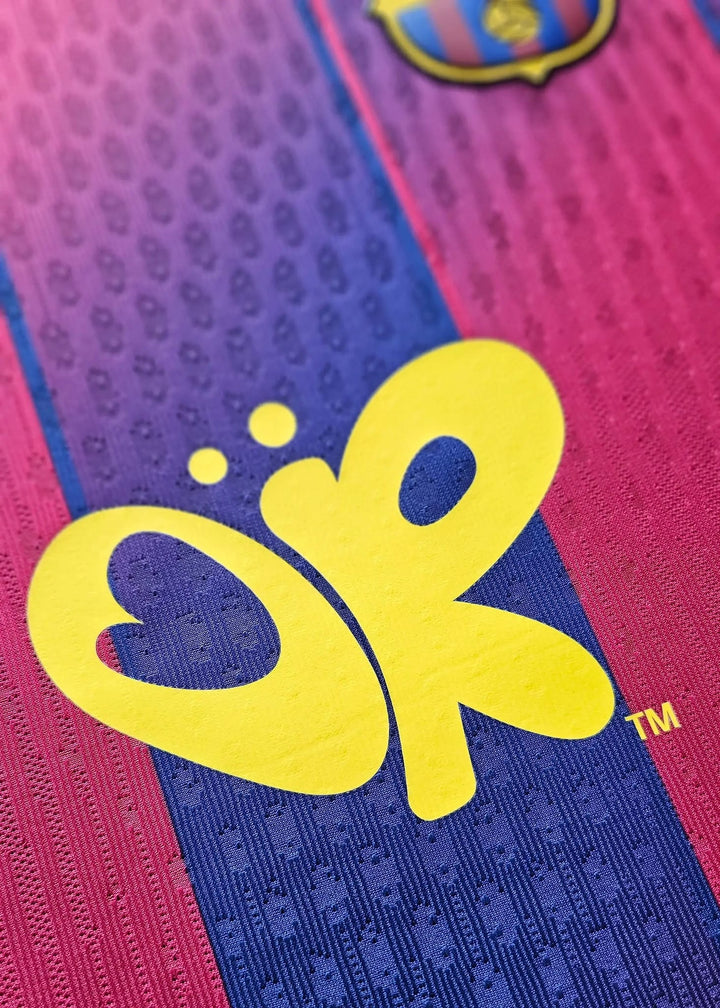A yellow butterfly 'OR' logo and the edge of the gold-and-blue Barcelona crest, on the maroon and navy 2025-2026 Olivia Rodrigo collaboration shirt.