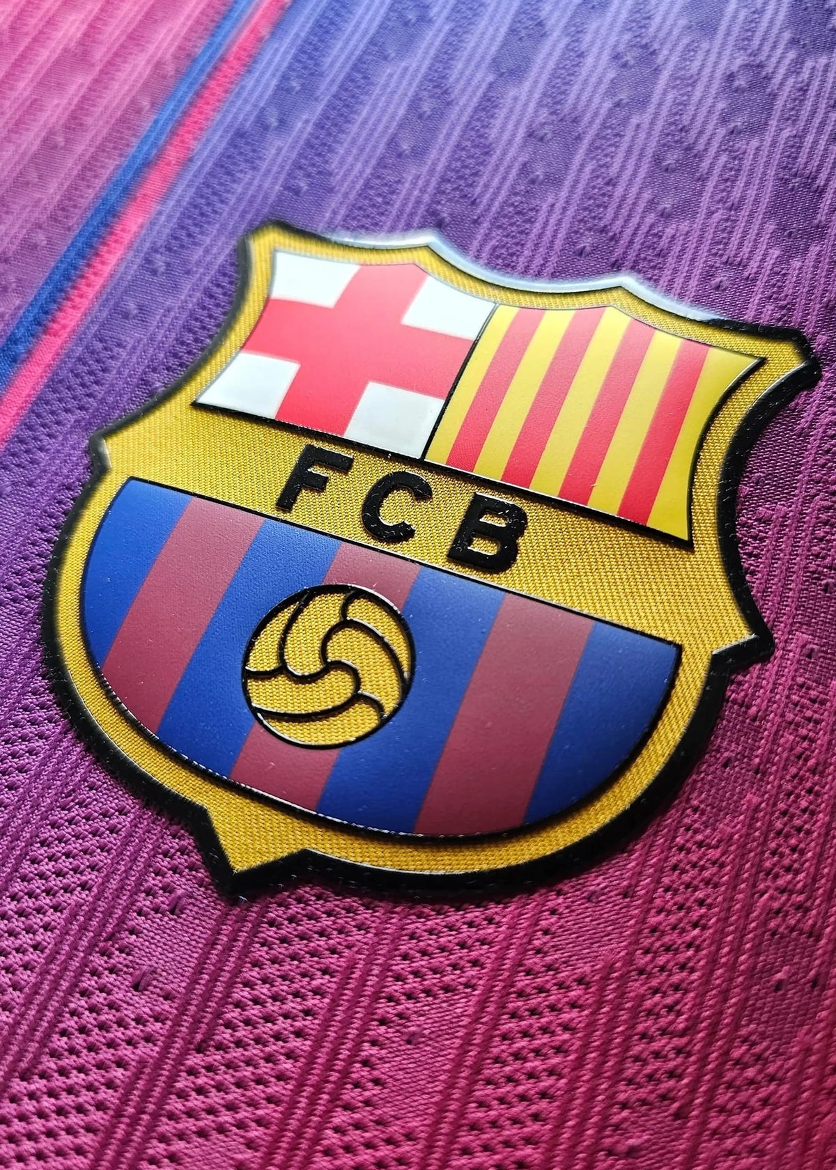 Close-up of the FCB crest on the purple and pink Barcelona Olivia Rodrigo collaboration shirt.