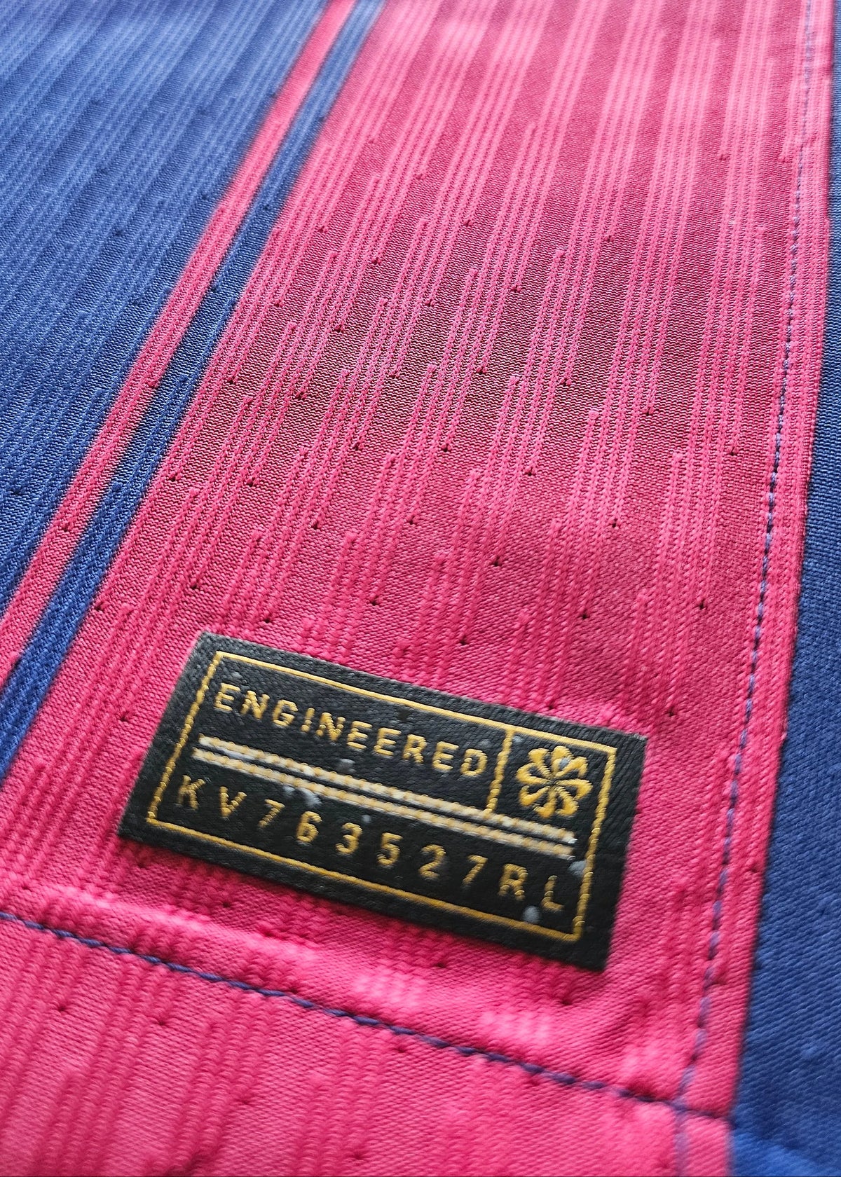 Close-up of a textured pink and blue striped fabric with a black and gold sewn-in label reading 'ENGINEERED' and 'KV763527RL'.