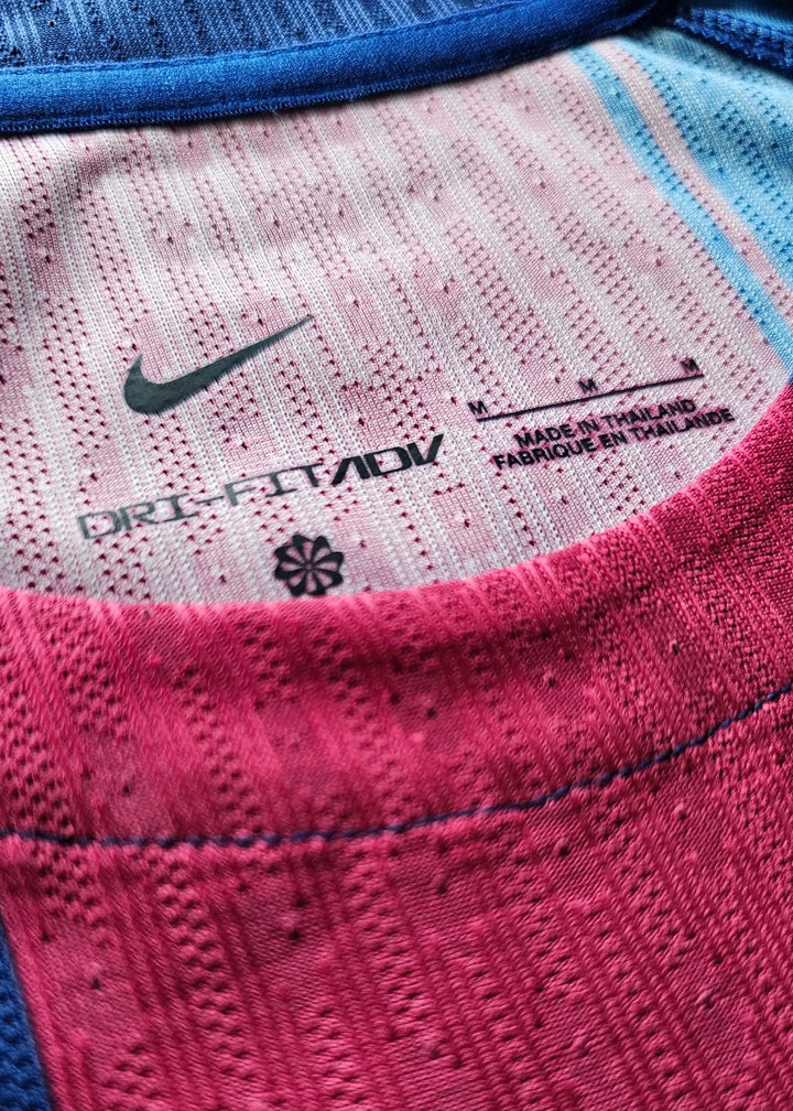Interior neck label with the Nike swoosh, 'DRI-FIT ADV', size M and 'Made in Thailand' text, on the pink-and-blue striped Barcelona shirt collar.