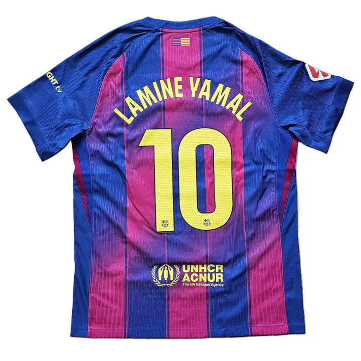 A FC Barcelona home jersey with the back of Lamine Yamal's name and number 10, laid flat on a light wood floor. The jersey features blue and magenta vertical stripes with yellow lettering and numbers.