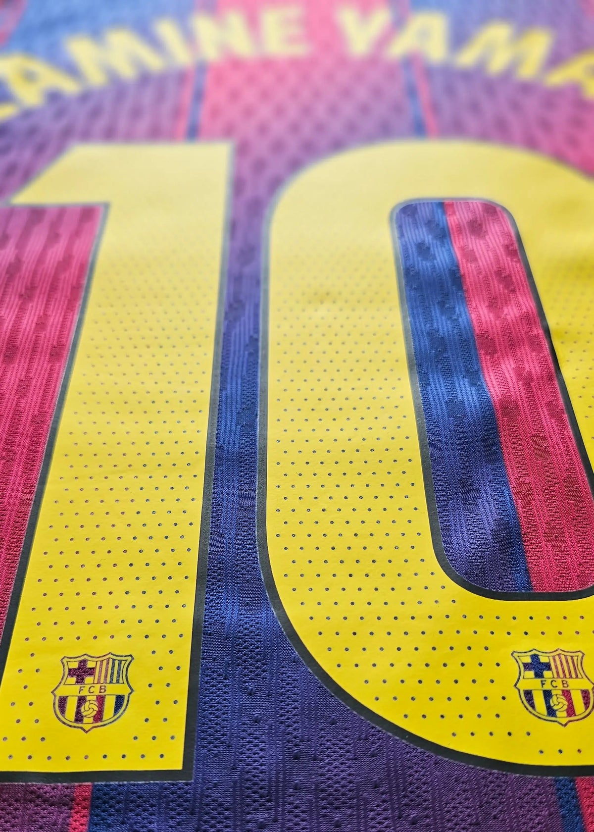 Close-up of the back of an FC Barcelona soccer jersey featuring the name YAMINE YAMAL and the number 10 in yellow, with the club crest near the bottom of the number. The jersey has a textured knit pattern in yellow, blue, and red.