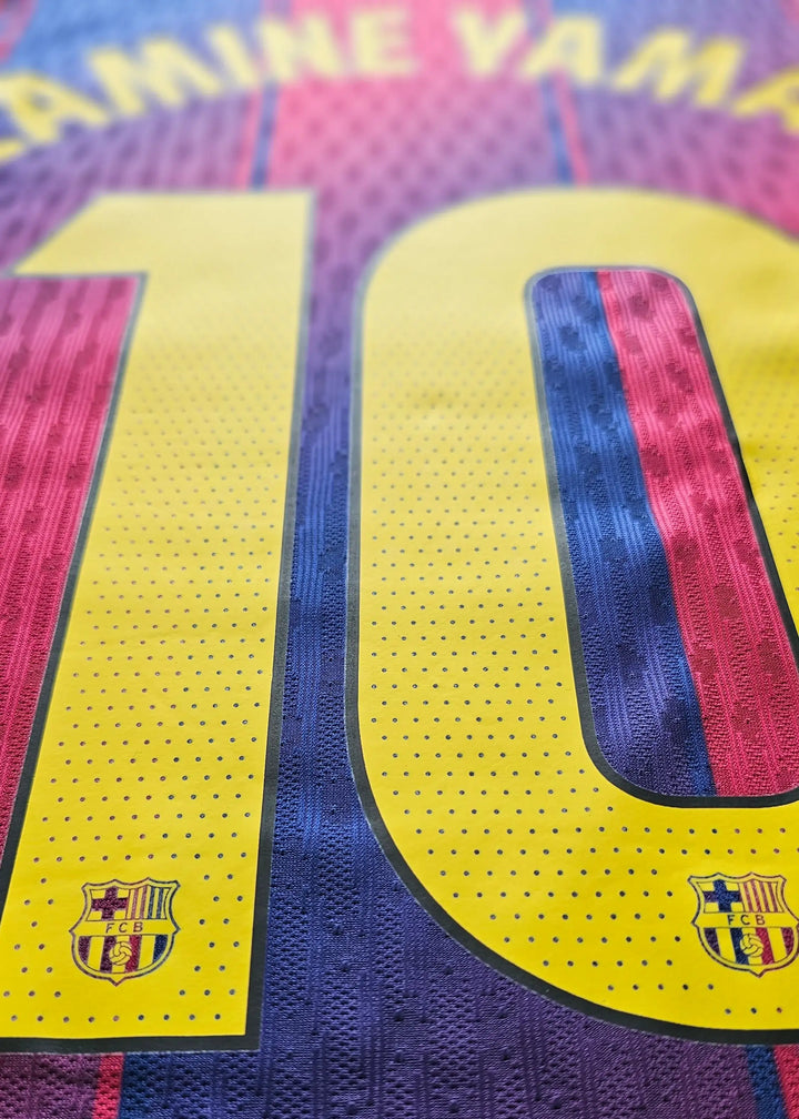 Close-up of the back of an FC Barcelona soccer jersey featuring the name YAMINE YAMAL and the number 10 in yellow, with the club crest near the bottom of the number. The jersey has a textured knit pattern in yellow, blue, and red.