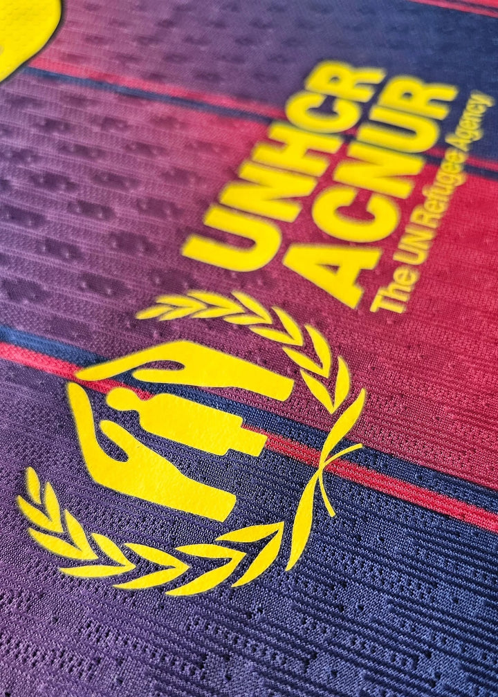 Close-up of a textured fabric surface with a large yellow UNHCR/ACNUR logo and the text 'The UN Refugee Agency' over a dark blue and red striped background.