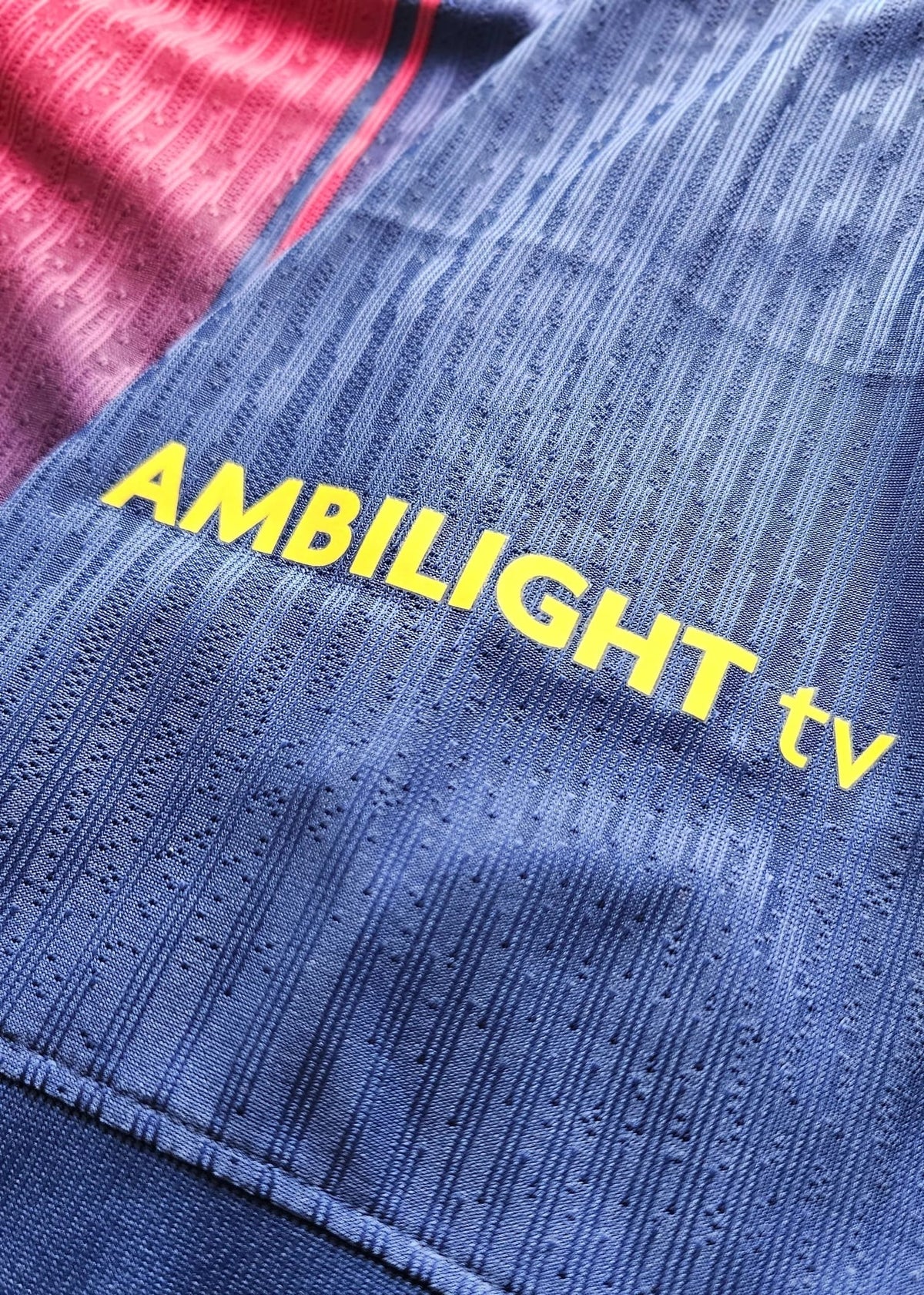 Close-up of a navy blue textured athletic jersey with yellow 'AMBILIGHT tv' text printed across the chest, with a red and navy second team jersey partially visible underneath.