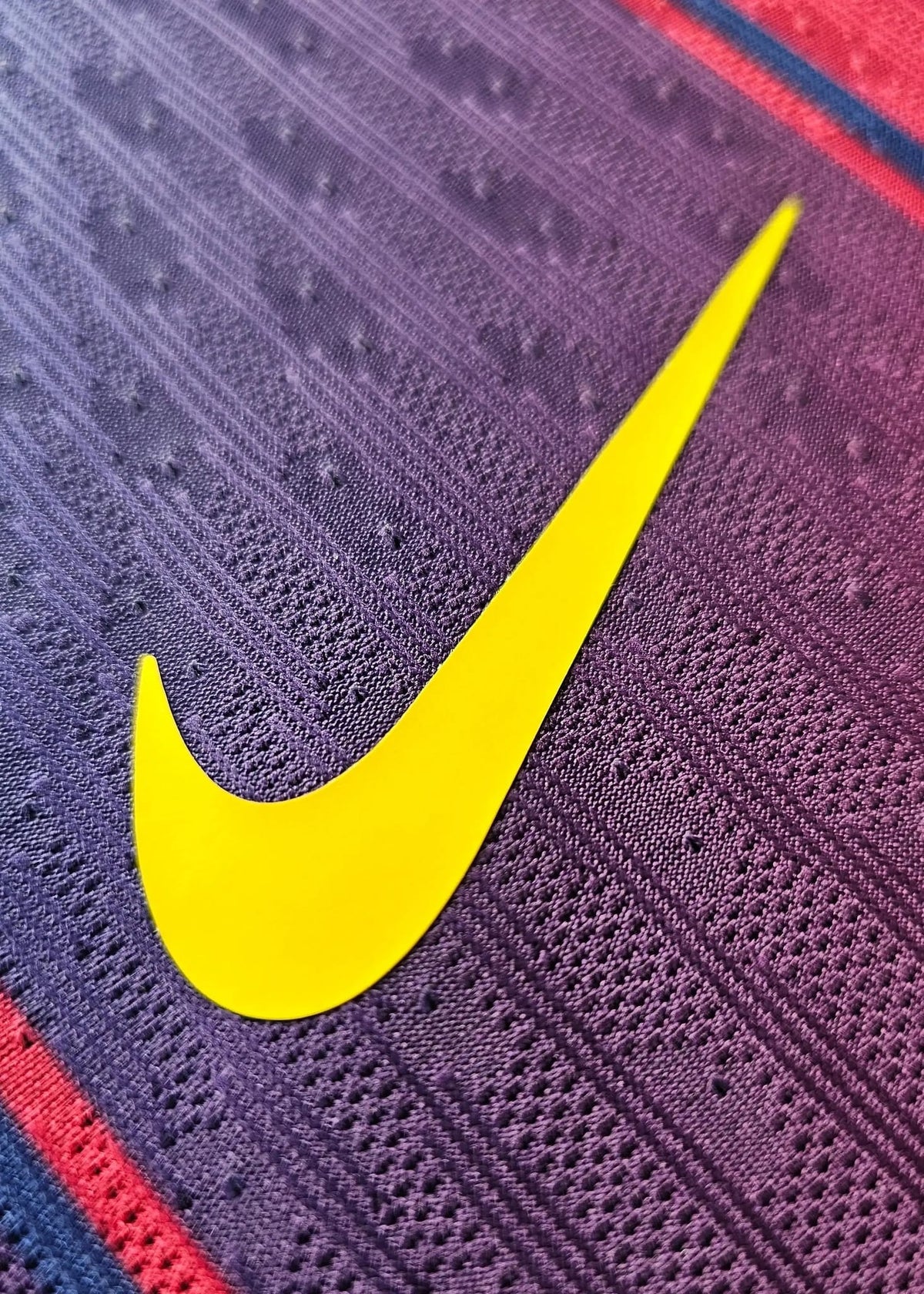 Close-up of a textured purple athletic fabric with a large bright yellow Nike swoosh logo and narrow red and navy stripes.