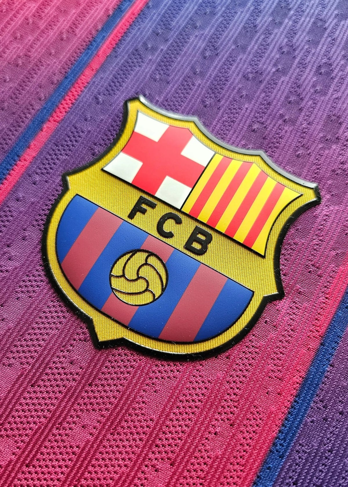 Full FC Barcelona crest with its red-and-white cross, yellow Catalan stripes, 'FCB' text and ball emblem, on the maroon-purple 2025-2026 shirt fabric.