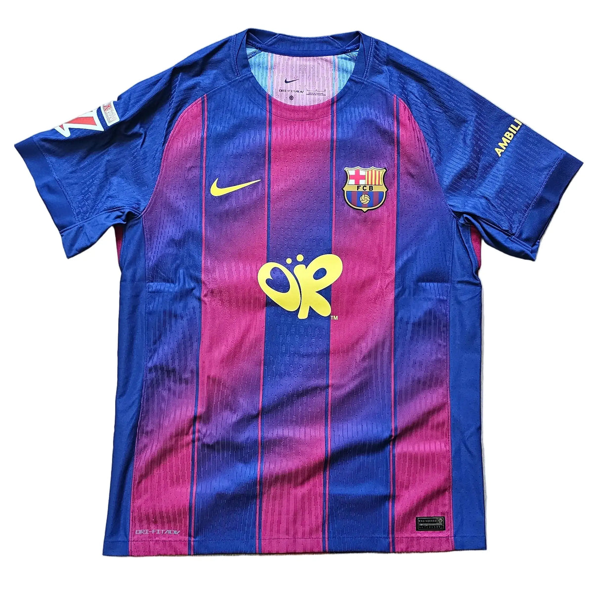 A Nike FC Barcelona home soccer jersey laid flat on a light wood floor. The jersey features the classic blue and magenta vertical stripes with a yellow gradient effect, a yellow Nike swoosh on the right chest, the FC Barcelona crest on the left chest, and a yellow sponsor logo in the center.