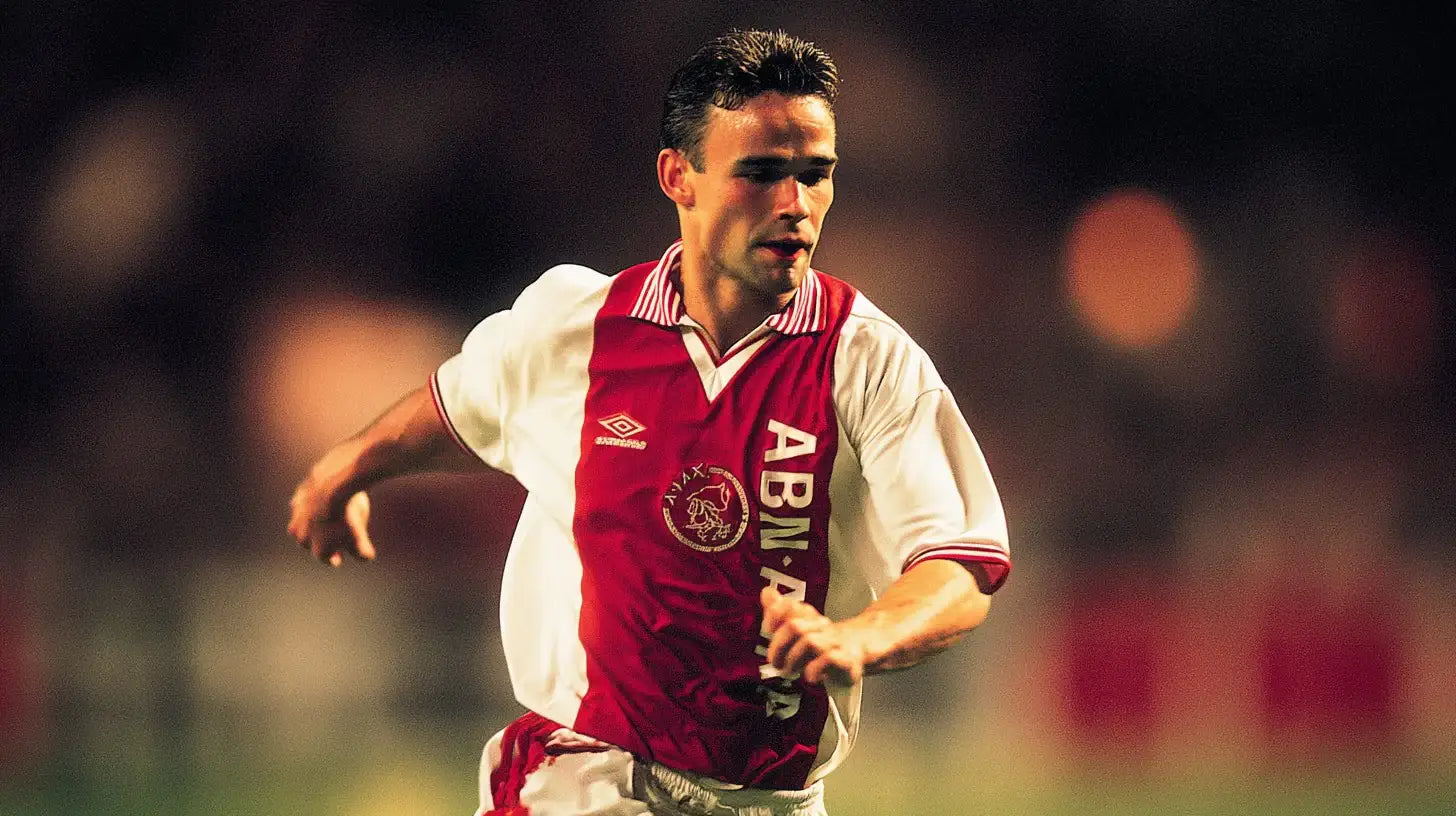 Marc Overmars running in an Ajax white football shirt with a red central panel and ABN AMRO sponsor, captured during a match with a soft-focus crowd.