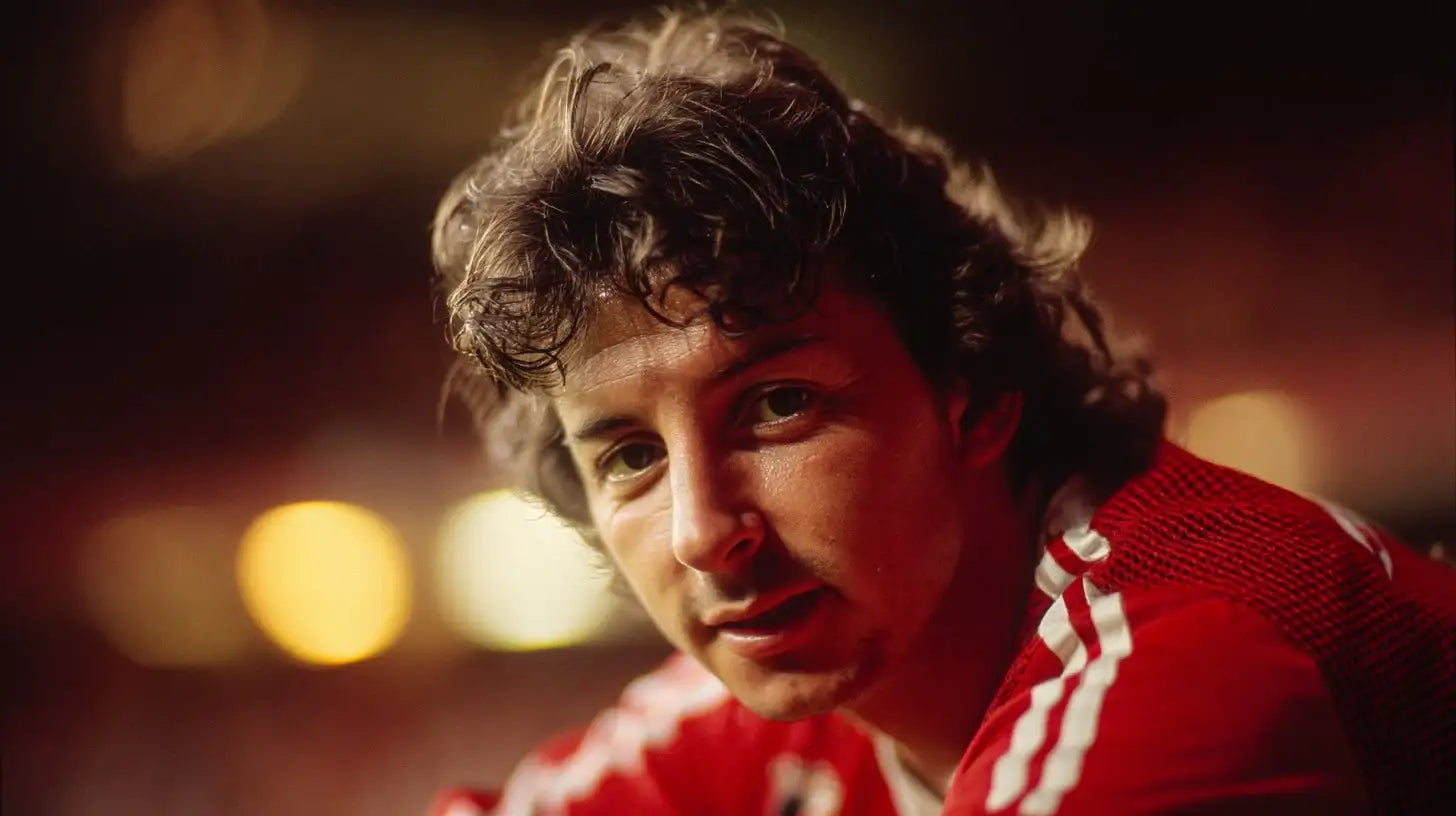 Mickey Thomas in a red Wrexham football shirt with white sleeve stripes, leaning forward with warm stadium lights out of focus behind him.