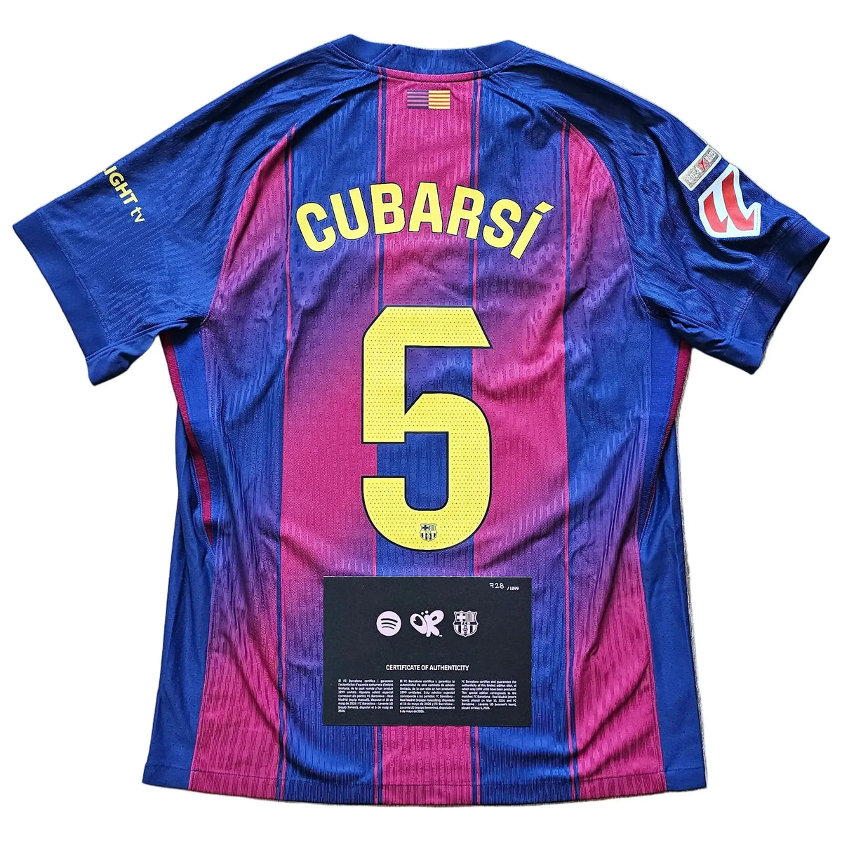 A FC Barcelona home soccer jersey with the back of the number 5 and player name CUBARSÁ visible, laid flat on a light wood floor. The jersey features blue and magenta vertical stripes with yellow lettering and number.