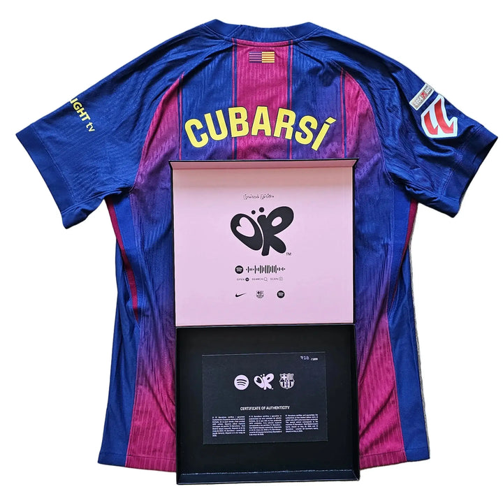 A blue and magenta striped FC Barcelona soccer jersey with 'CUBARSI' printed in yellow on the back, laid flat on a light wood floor with a framed award plaque placed in front of it.