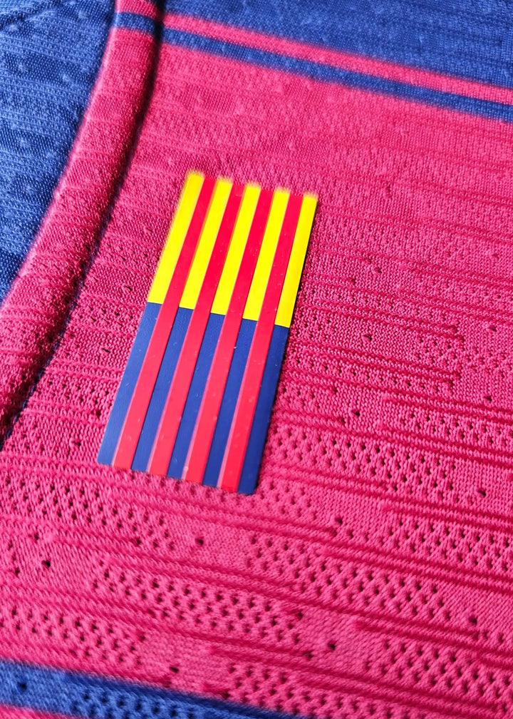 Close-up of red-yellow Barça crest stripes stitched onto the maroon-and-blue knit of Pau Cubarsí's FC Barcelona 2025-2026 shirt.