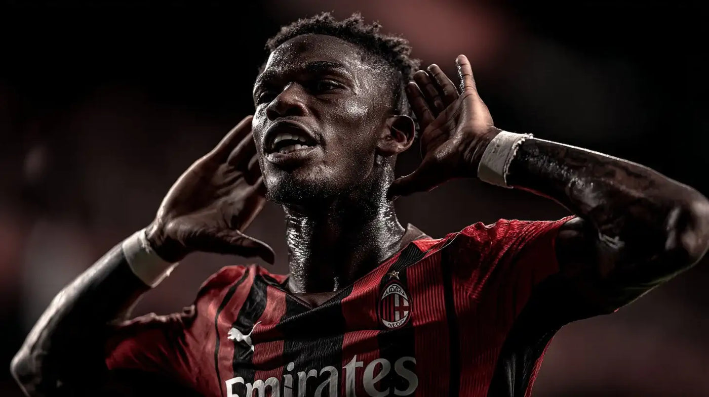 Rafael Leão shouts toward the crowd in an AC Milan black and red striped home football shirt, hands to his ears under dramatic dark stadium lights.
