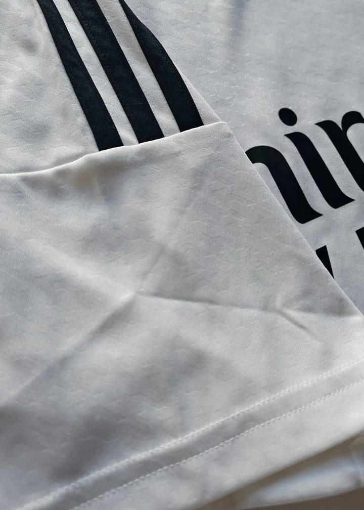 This detail shot shows the three black stripes along the sleeve of the Raul Asencio Real Madrid 2024-2025 shirt, with partial print lettering visible beside it.
