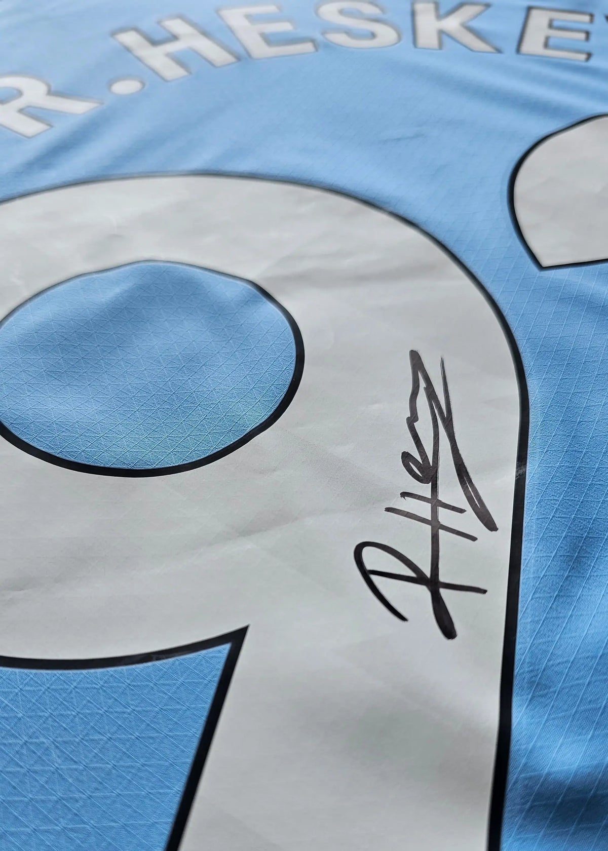 Close-up of a black signature beside the white No. 9 in HESKEY on a sky-blue Manchester City shirt, signed by Reigan Heskey.