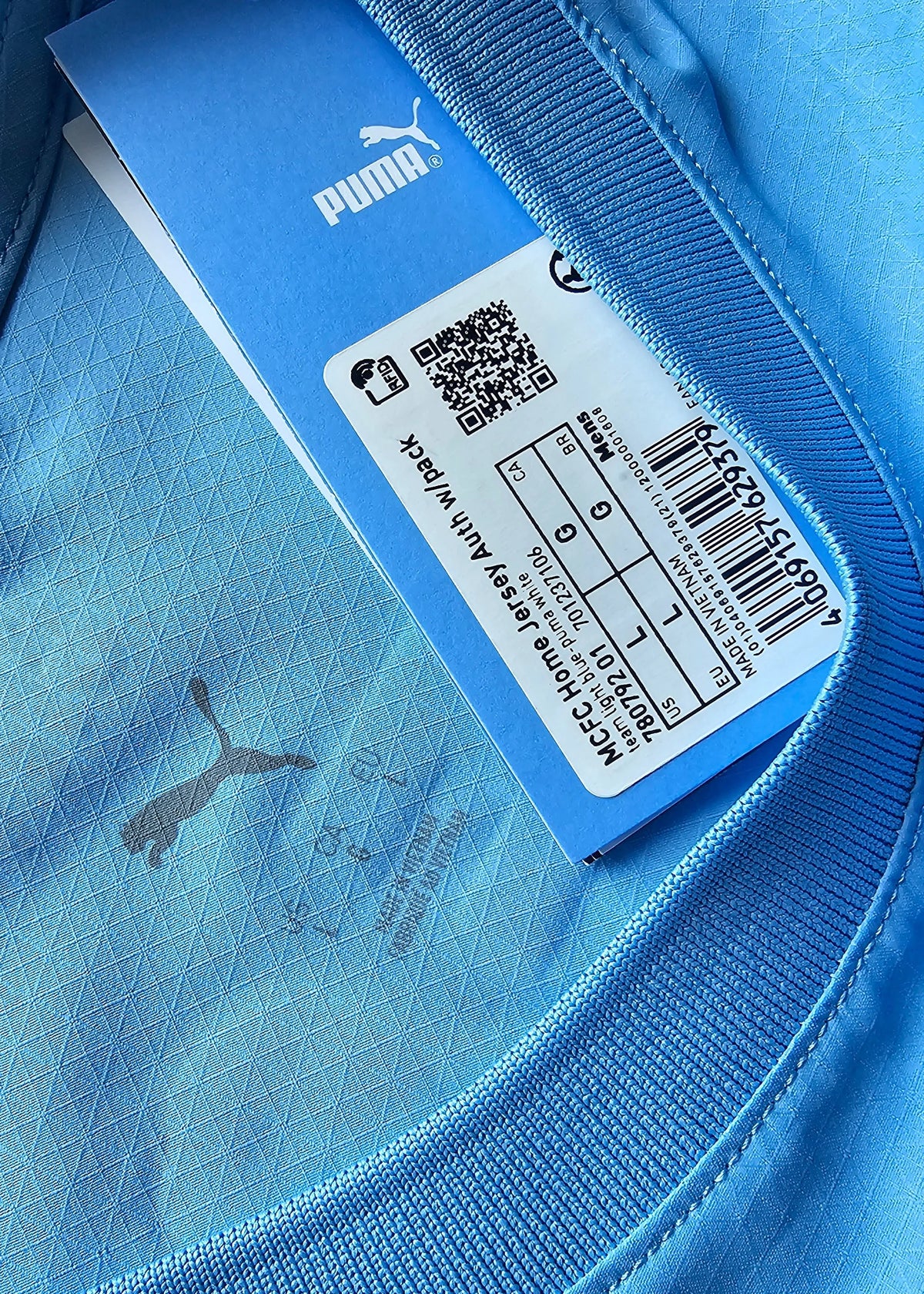 Close-up, shown upside down, of a Puma swing tag reading 'MCFC Home Jersey' with a size label and barcode, on the signed Reigan Heskey shirt.