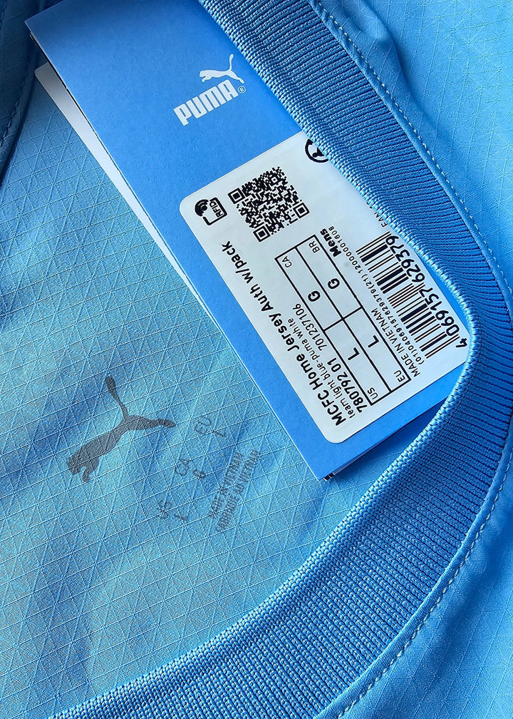 Close-up, shown upside down, of a Puma swing tag reading 'MCFC Home Jersey' with a size label and barcode, on the signed Reigan Heskey shirt.