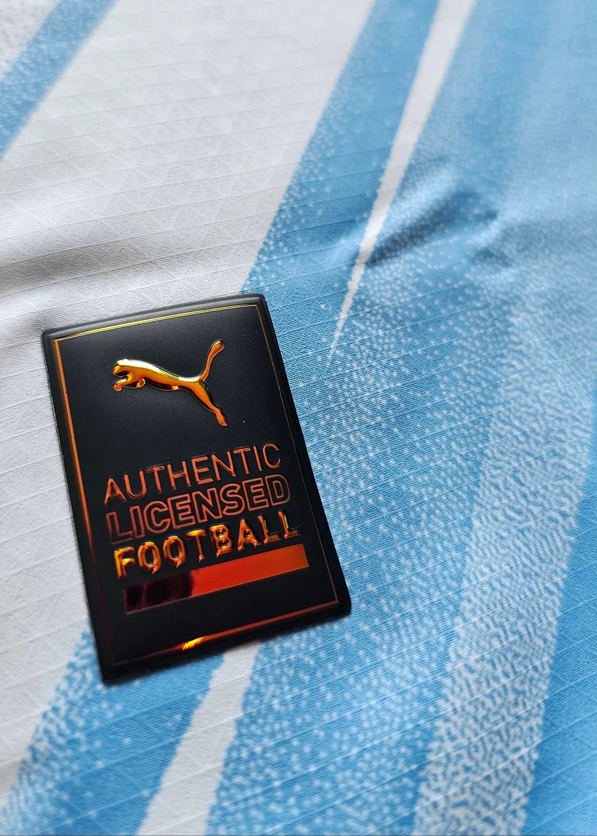 Close-up of a black Puma 'Authentic Licensed Football' tag on white-and-sky-blue striped fabric, from the signed Reigan Heskey Man City shirt.