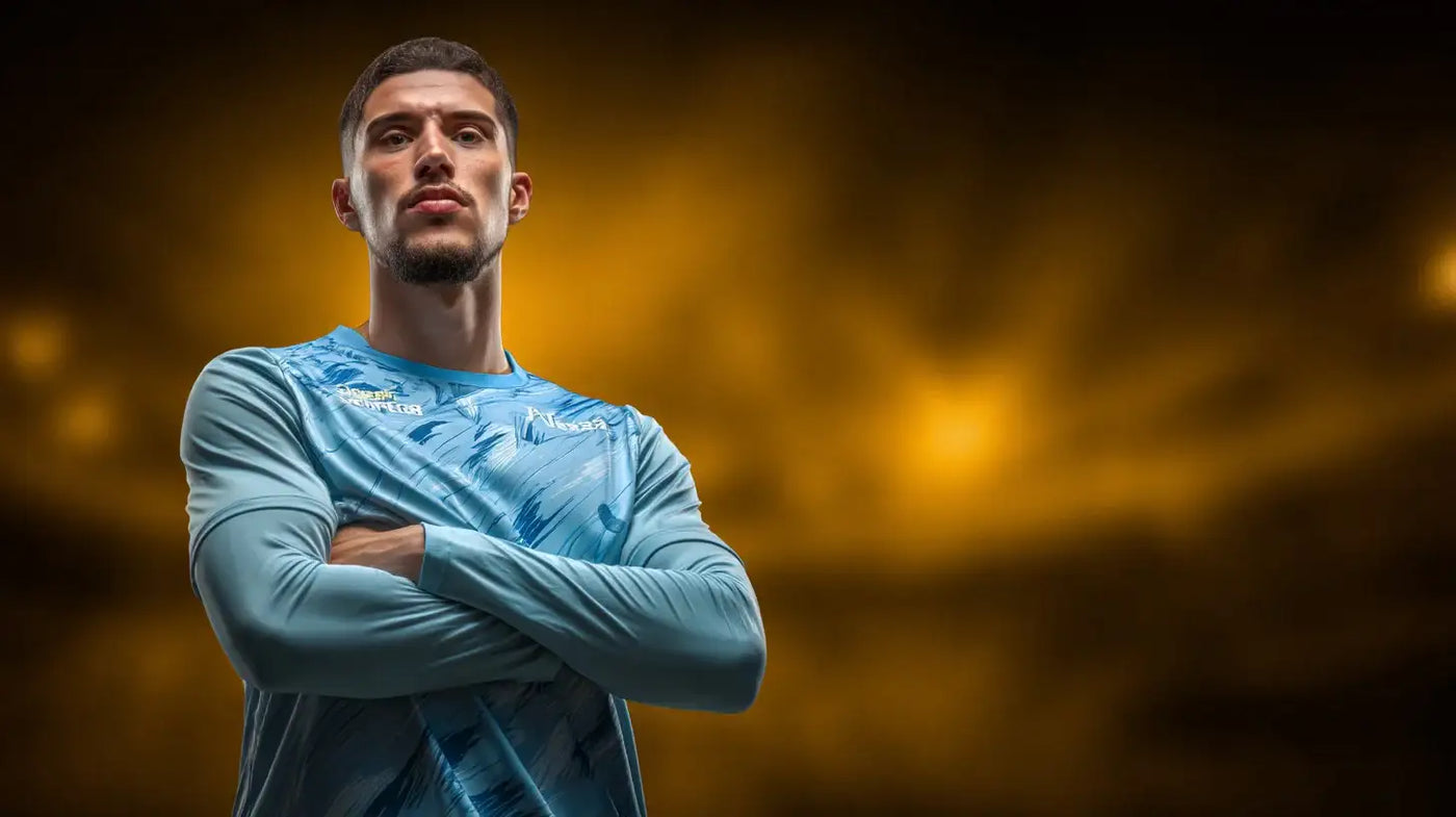 Robin Risser standing with arms crossed in a light blue Lens football kit, warm stadium lights blurred behind him.