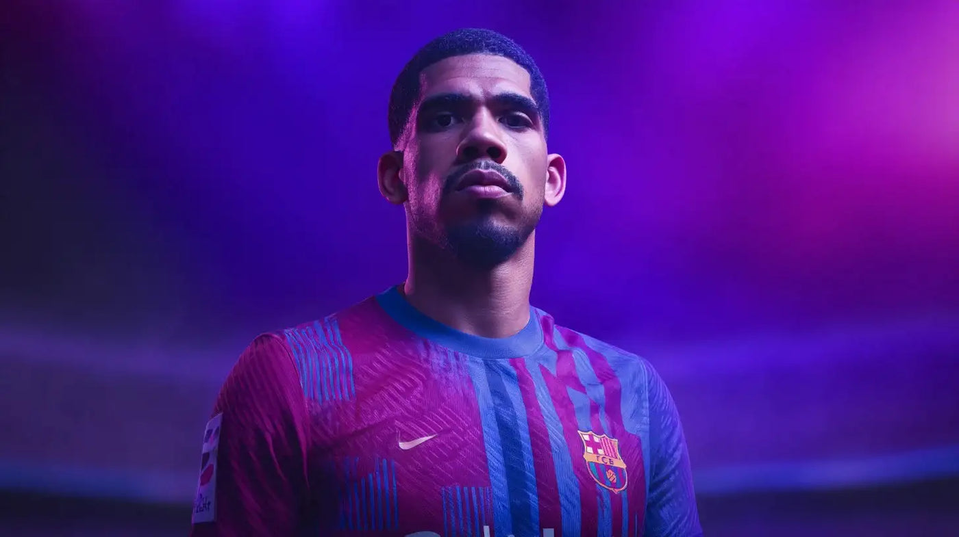 Ronald Araújo in a Barcelona home football shirt with blue and red vertical stripes stands facing the camera with a serious expression, lit by strong purple and blue stadium lights.