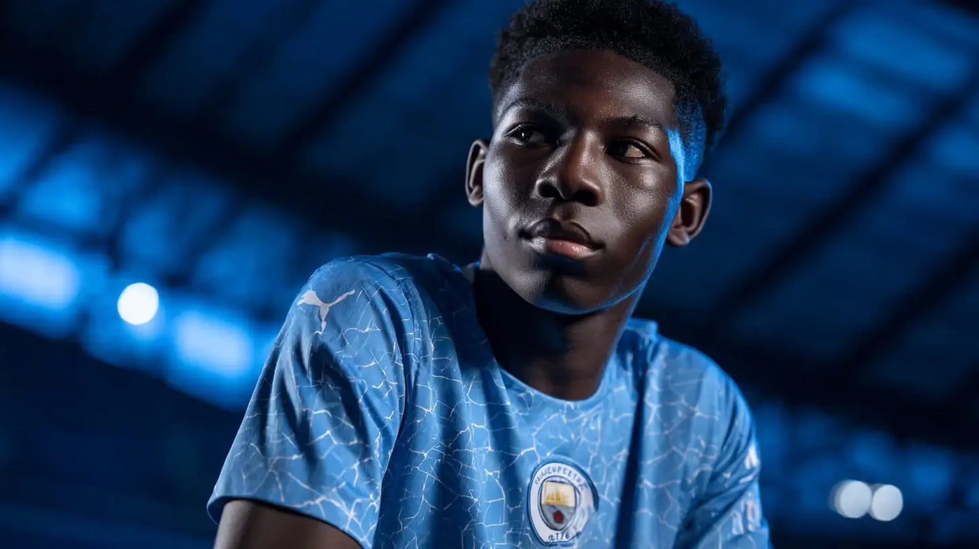 Ryan McAidoo in Manchester City sky-blue shirt, close-up portrait looking right with blurred stadium background.