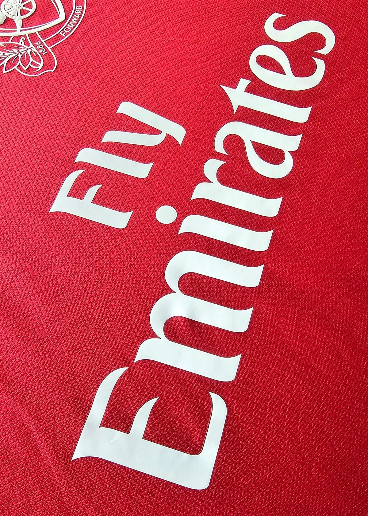 Close-up of the white Fly Emirates wordmark printed diagonally on a red Arsenal shirt, with the Arsenal crest visible in the corner, No. 10.
