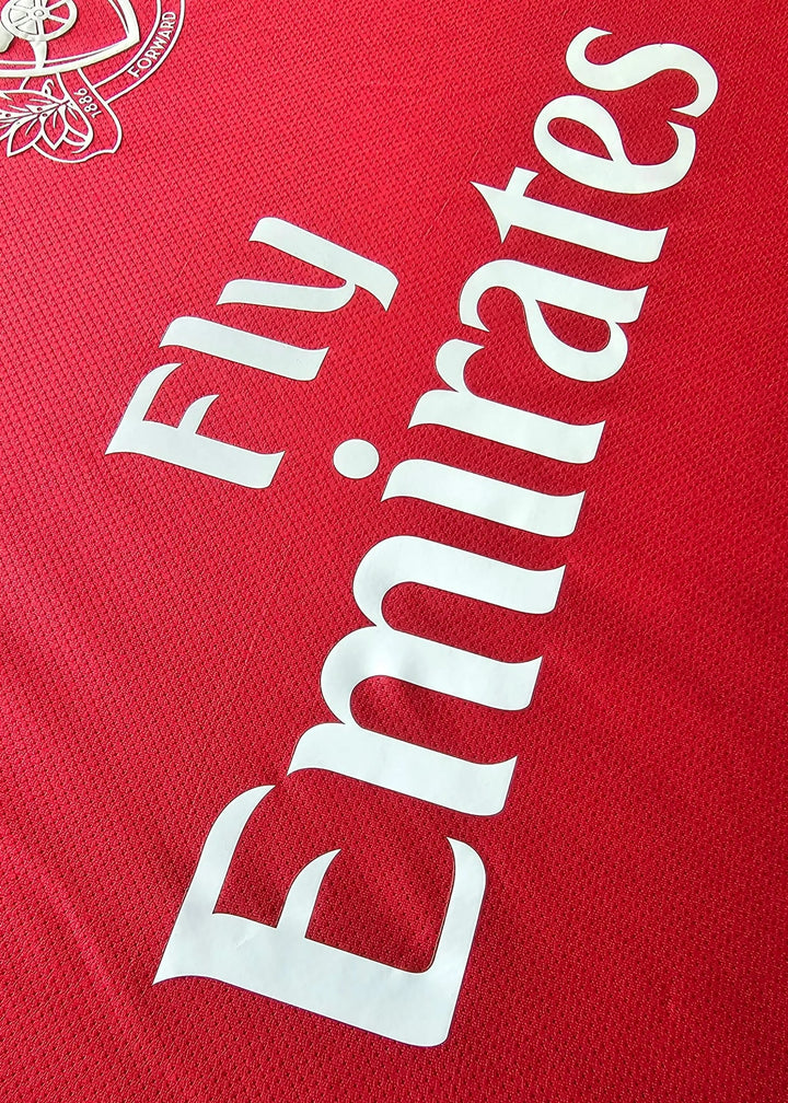 Close-up of the white Fly Emirates wordmark printed diagonally on a red Arsenal shirt, with the Arsenal crest visible in the corner, No. 10.