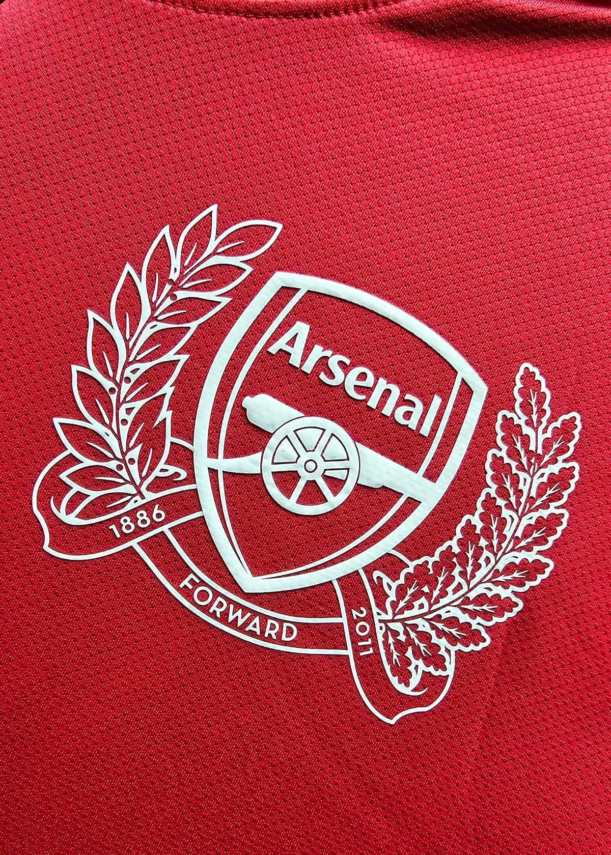 Close-up of the white Arsenal crest with cannon, wreath and 1886-2011 banding, printed on the chest of a red 2011-12 No. 10 shirt.