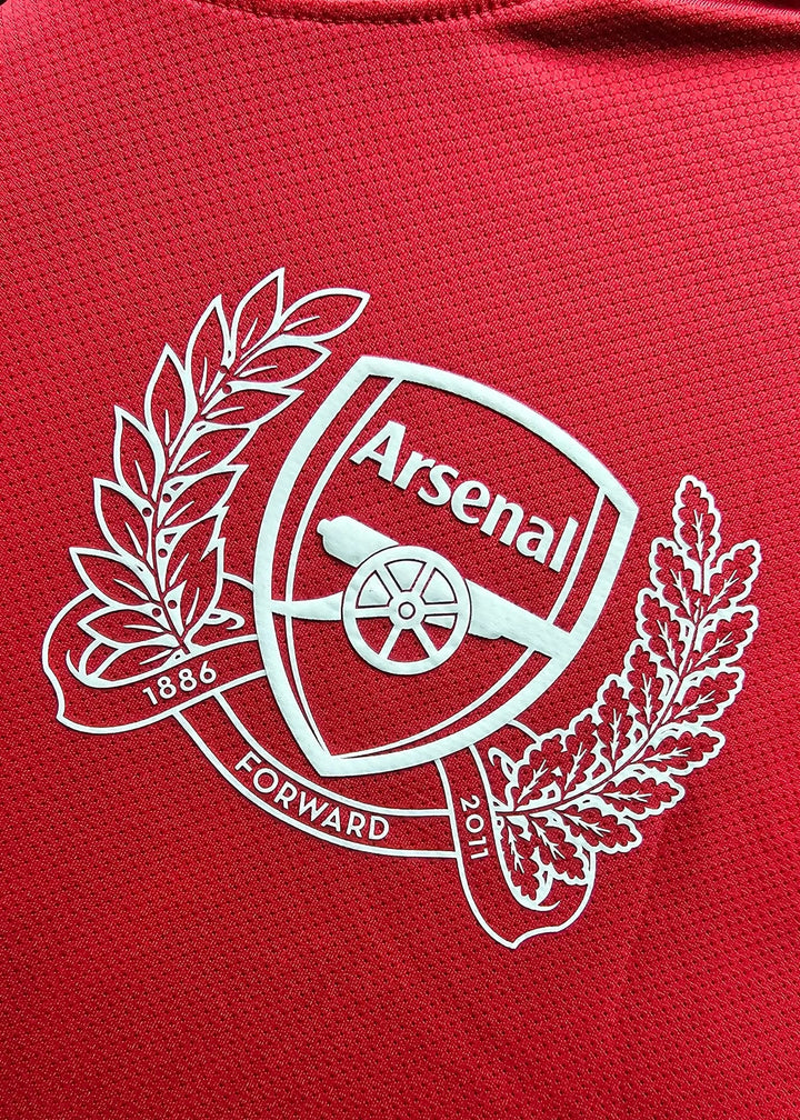 Close-up of the white Arsenal crest with cannon, wreath and 1886-2011 banding, printed on the chest of a red 2011-12 No. 10 shirt.