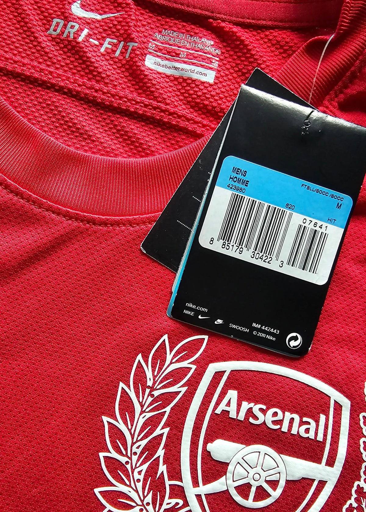 Interior collar tag reading DRI-FIT with the Nike swoosh, plus an attached retail swing tag with a barcode, Mens Homme M, beside the Arsenal crest.