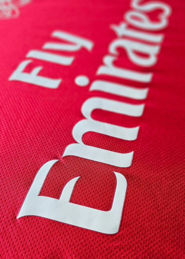 Fly Emirates sponsor lettering printed on red fabric, from Robin van Persie's signed Arsenal 2011-12 Champions League No. 10 shirt front.