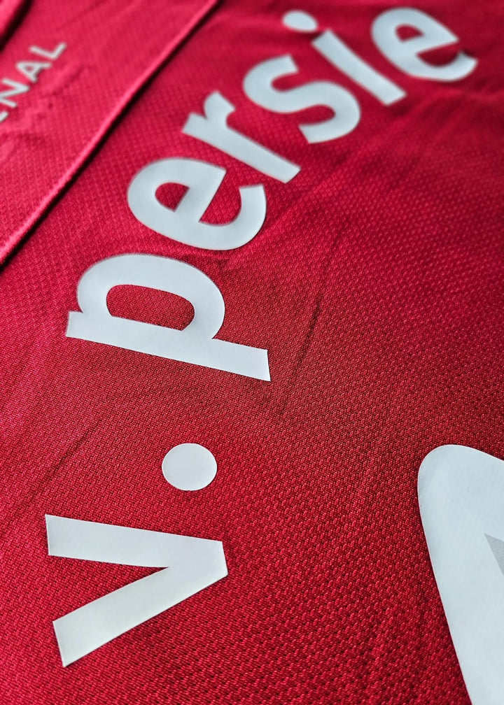 Close-up, diagonal view of the white v. persie script nameplate, with ARSENAL lettering visible in the corner, on the Champions League shirt.